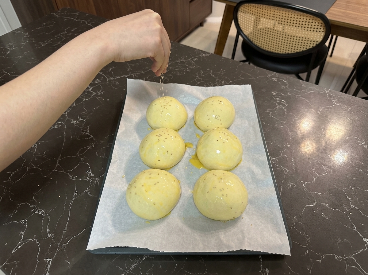 Una mano che sparge minuscoli semi di sesamo bianchi sulle palline di impasto spennellate duovo disposte su una teglia.