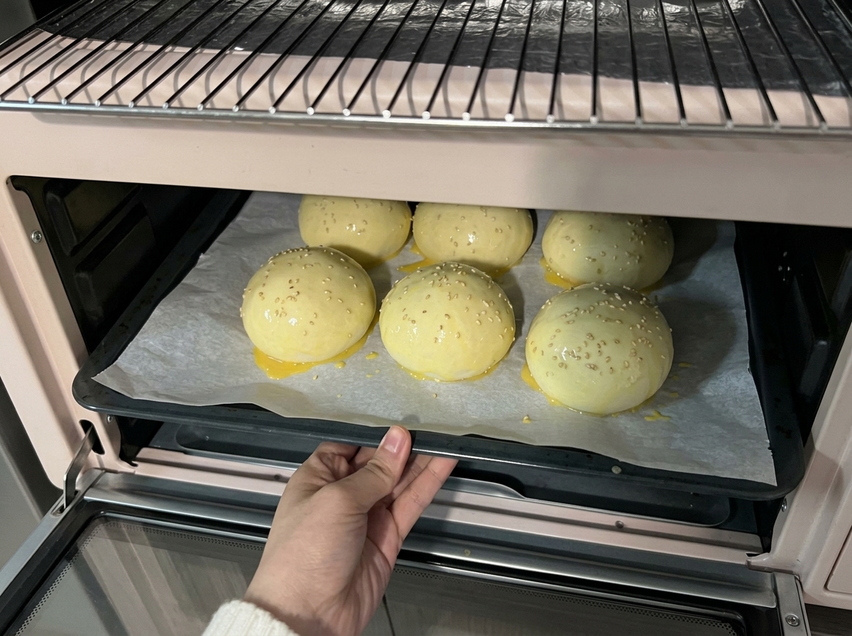 Una teglia di palline di impasto ricoperte di sesamo infilata in un piccolo forno rosa chiaro per la cottura.
