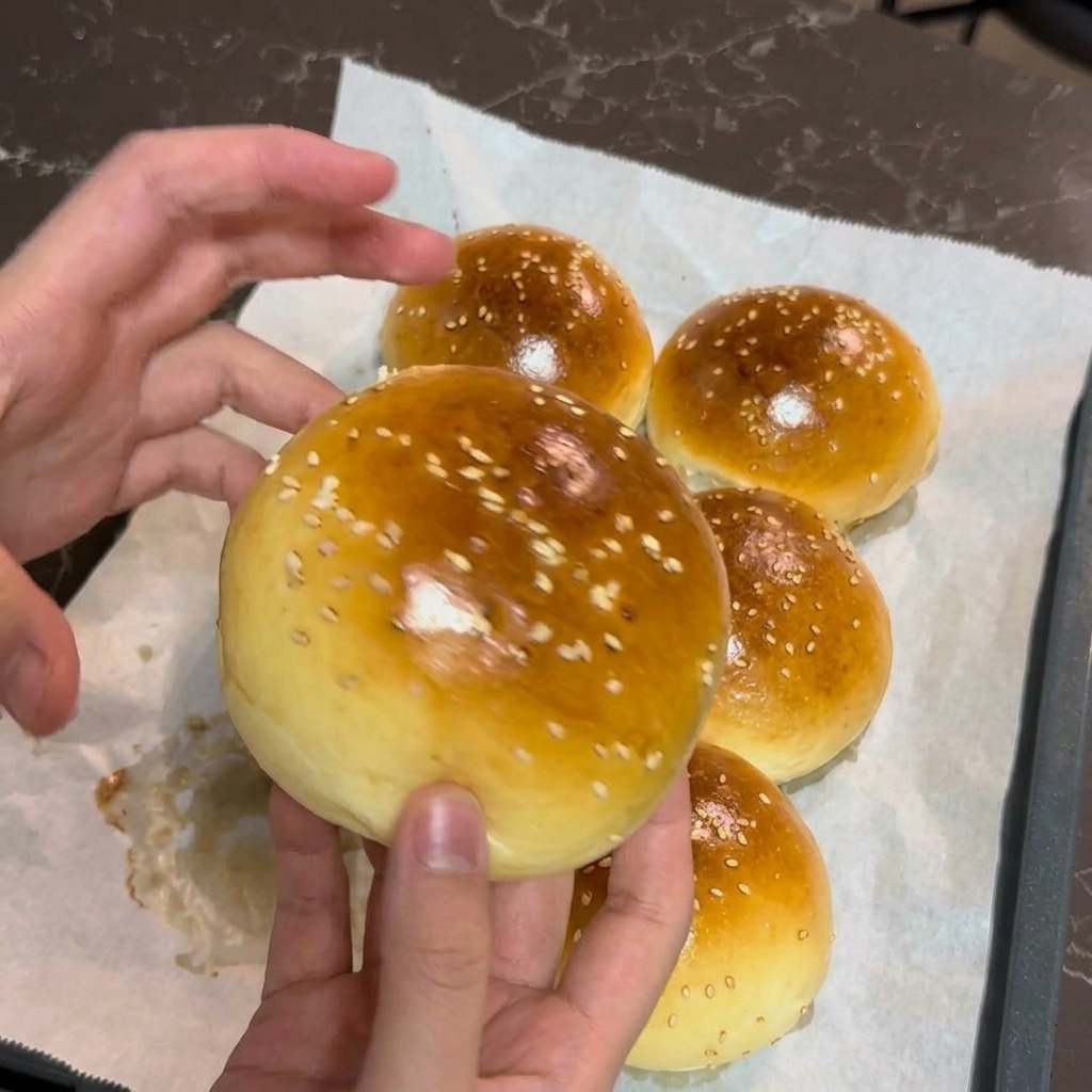Le mani di una persona che tengono delicatamente un panino da hamburger appena sfornato, dorato e cosparso di semi di sesamo bianchi.
