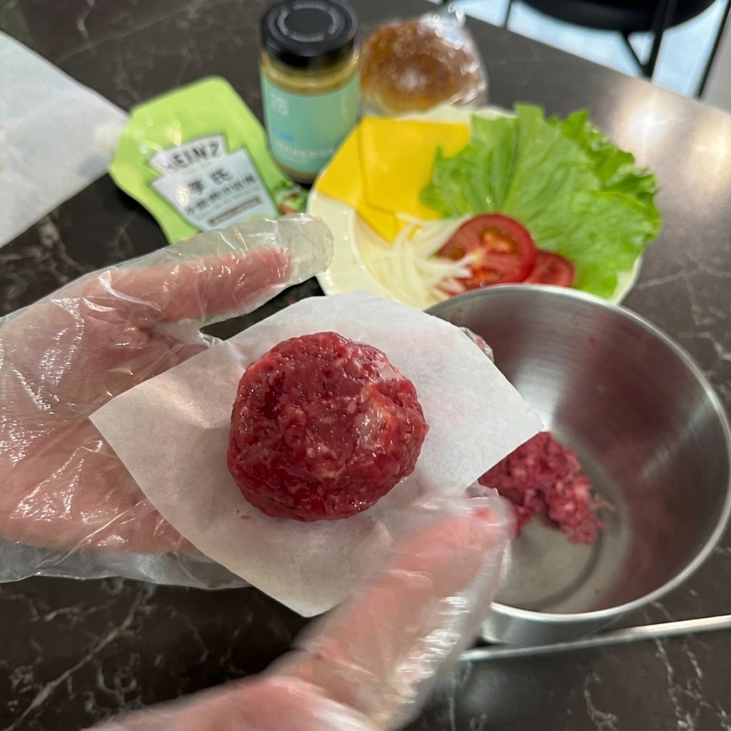 Una mano guantata che tiene una palla rotonda di carne di manzo cruda poggiata su un quadratino di carta da forno bianca.