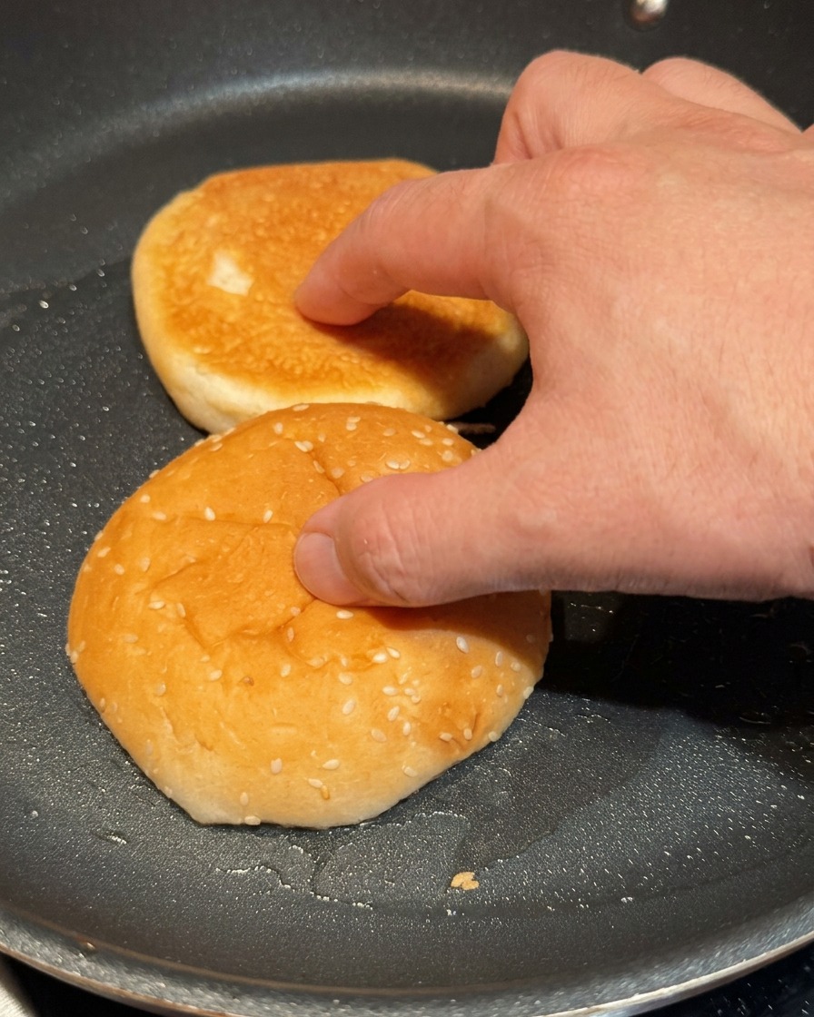 Una mano che preme leggermente le due metà di un panino per hamburger su una padella calda per tostarle.