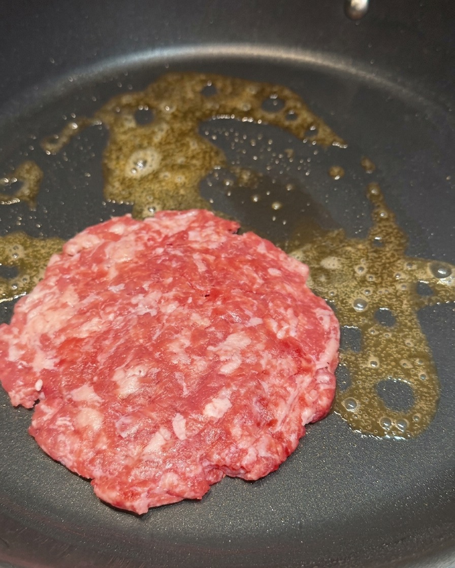 Un hamburger di manzo crudo e marmorizzato che sfrigola nel burro fuso in padella.