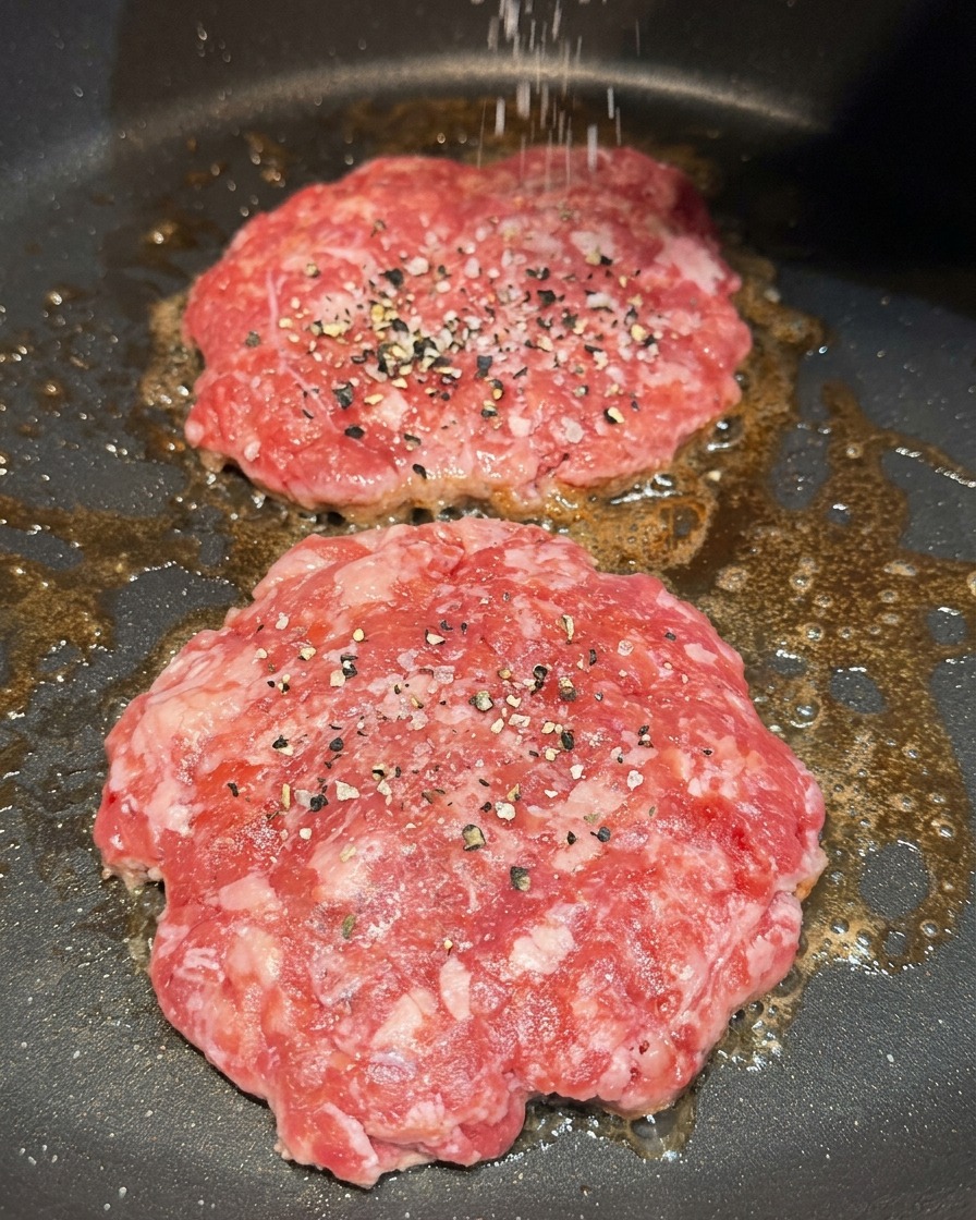 Cospargere sale grosso e pepe nero macinato su due hamburger crudi che cuociono in padella.