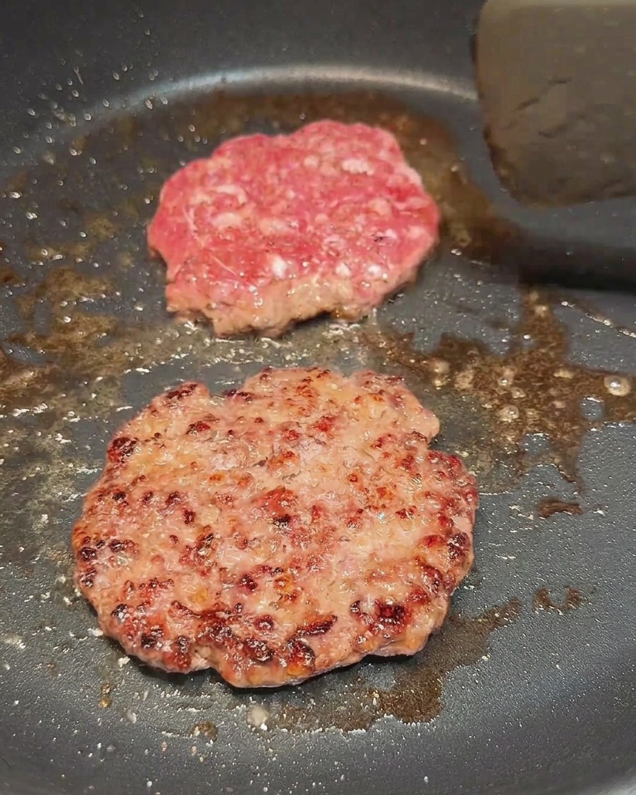 Due hamburger di manzo in padella, di cui uno appena girato che mostra una crosticina ben dorata.