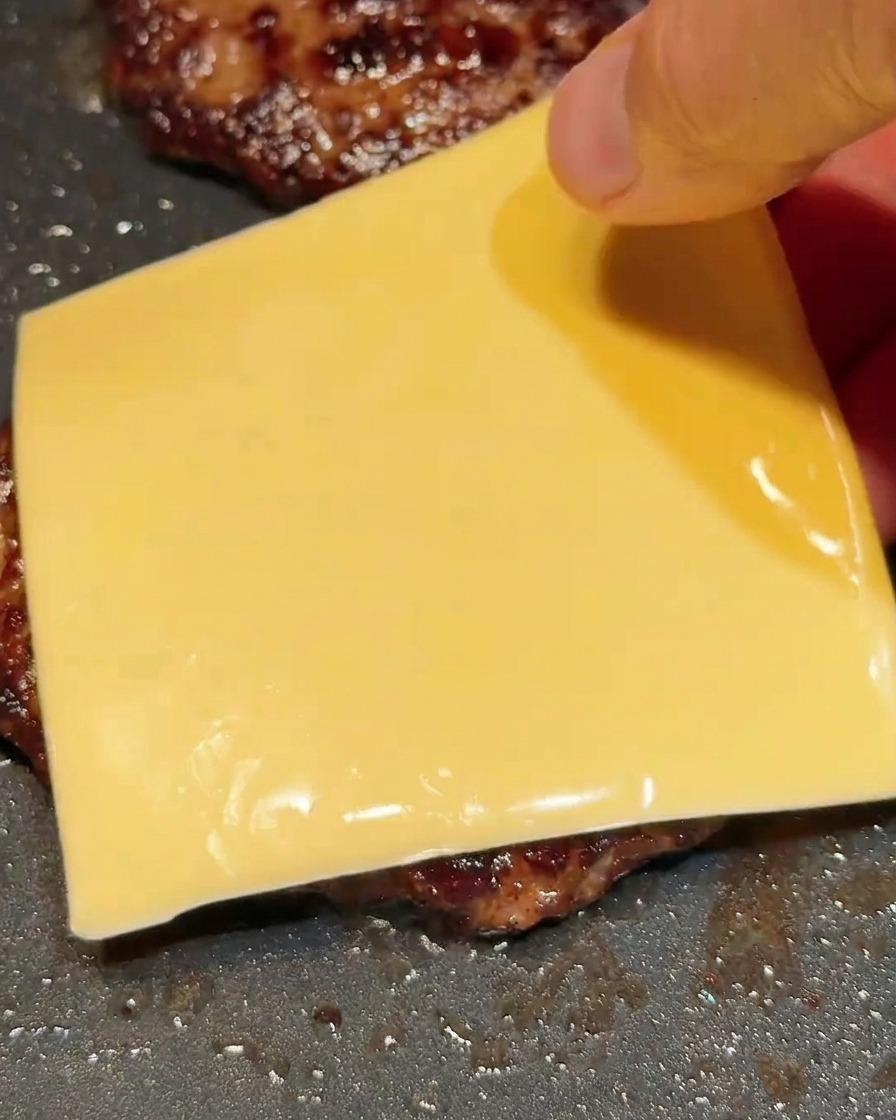 Una mano che adagia una fetta quadrata di formaggio su un hamburger caldo in padella.