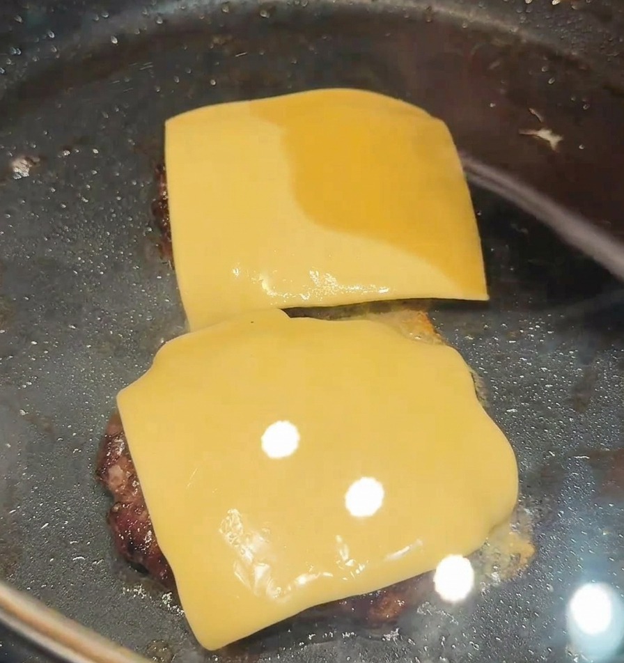Due fette quadrate di formaggio posate sopra due hamburger che sfrigolano in padella.