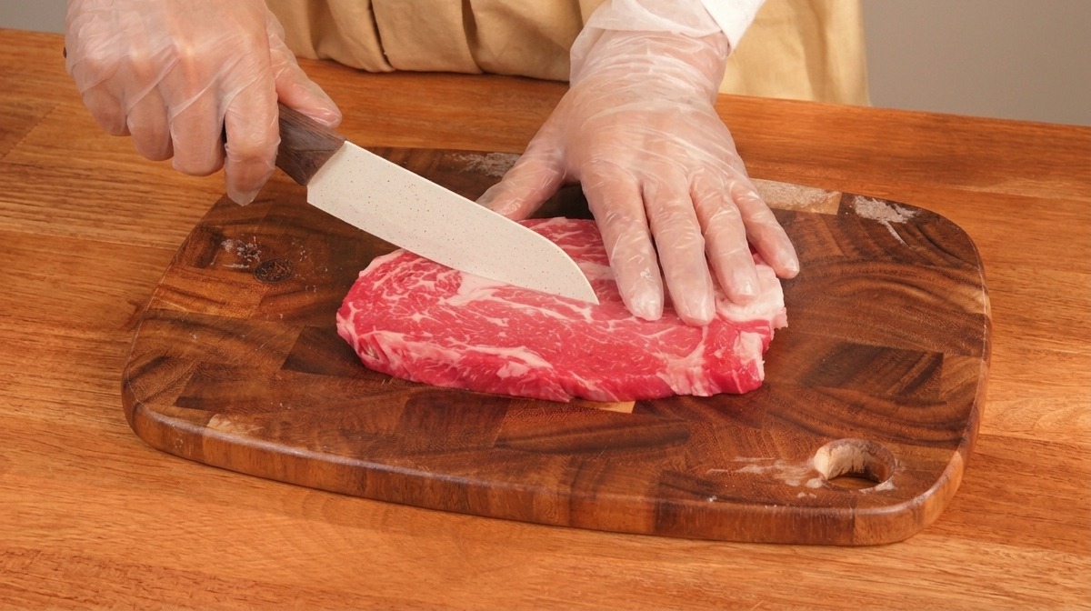 Mani che tagliano a cubetti un pezzo di manzo marezzato su un tagliere di legno.
