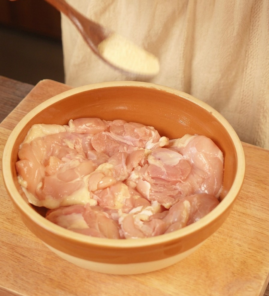 Un cucchiaio di legno che sparge aglio in polvere su sovracosce di pollo crudo in una ciotola di ceramica marrone.