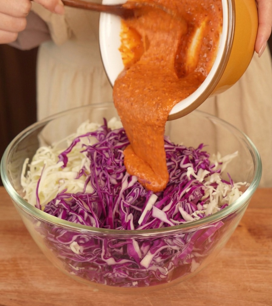 Dressing arancione versato da una ciotola gialla su una montagna di cavolo bianco e viola tritato in una ciotola di vetro.