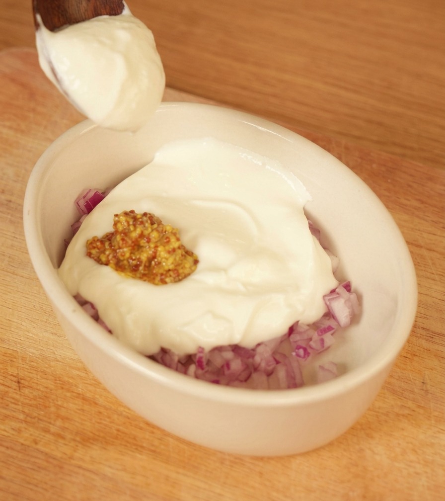 Un cucchiaio di yogurt bianco denso aggiunto a una ciotolina con cipolle rosse tritate e senape.