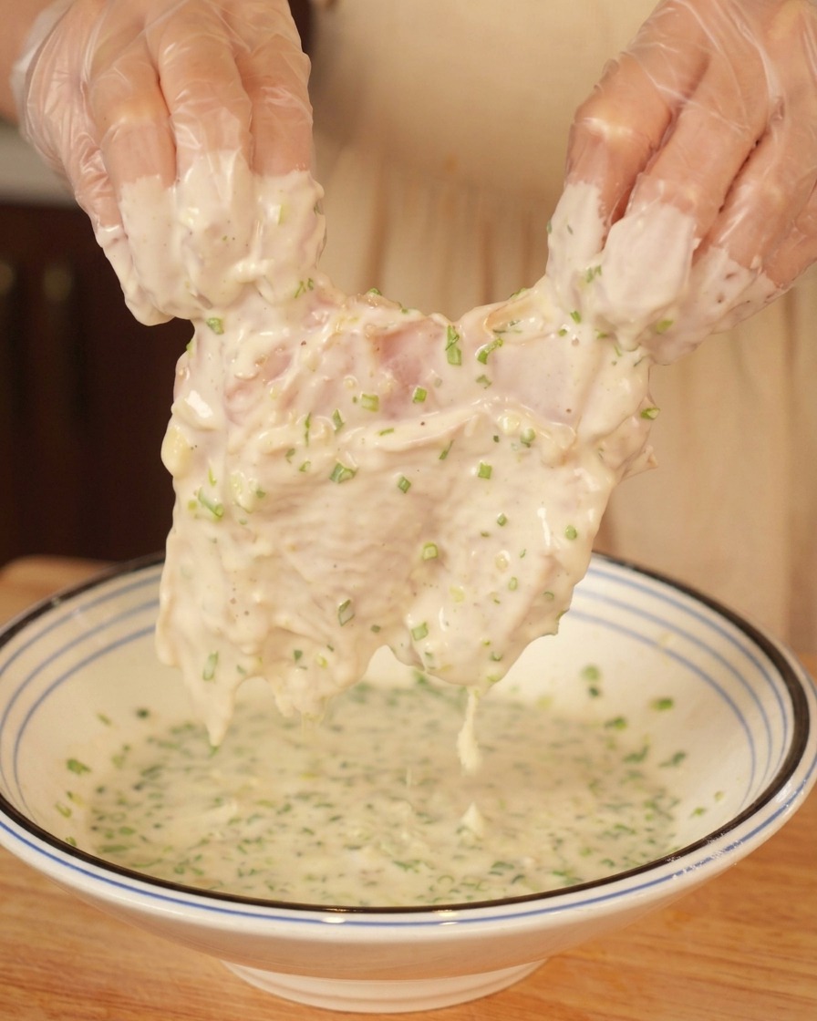 Mani guantate che sollevano una sovracoscia di pollo ricoperta da una densa pastella bianca punteggiata di verde.