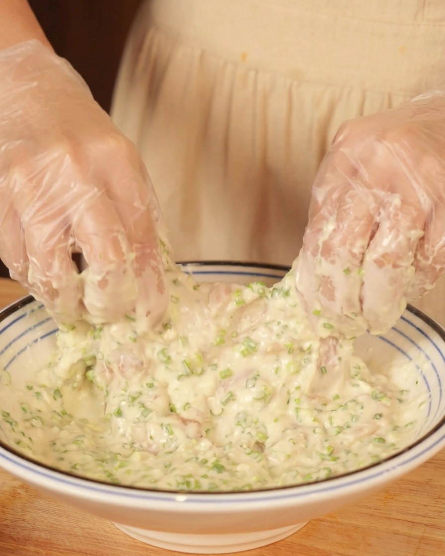 Mani guantate che ricoprono un pezzo di pollo crudo in una ciotola di pastella densa.