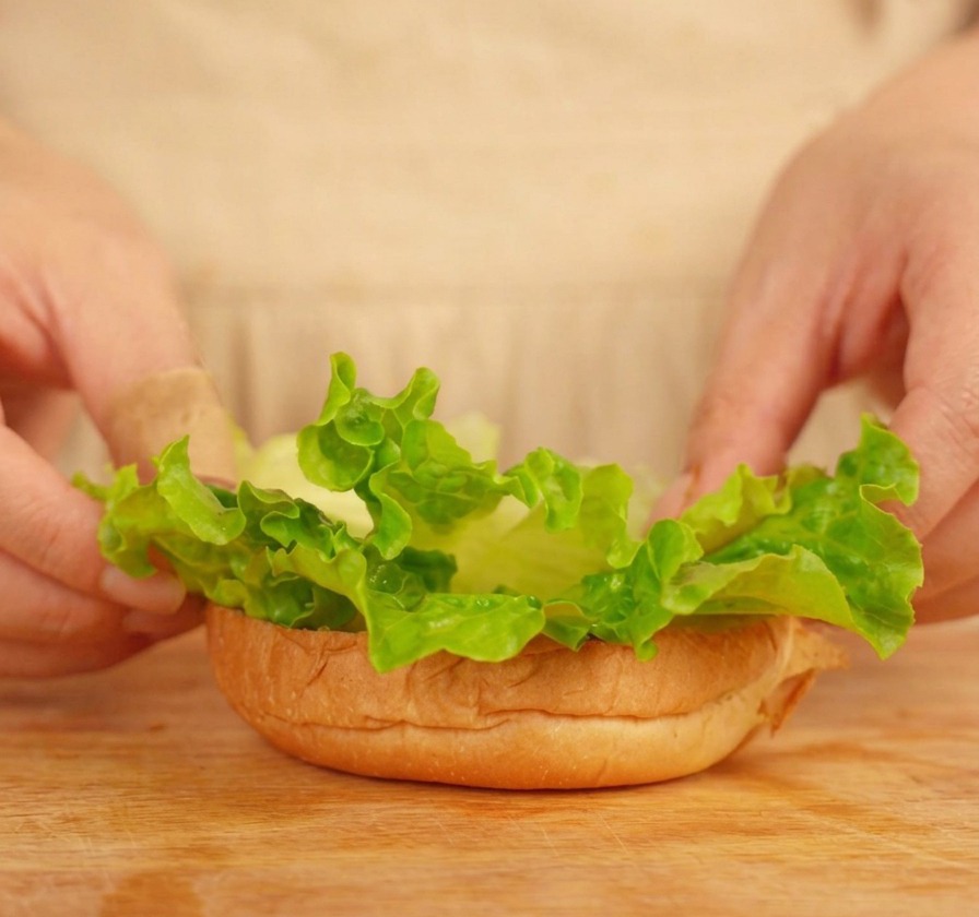 Mani che posizionano foglie di lattuga sulla base di un panino tostato.