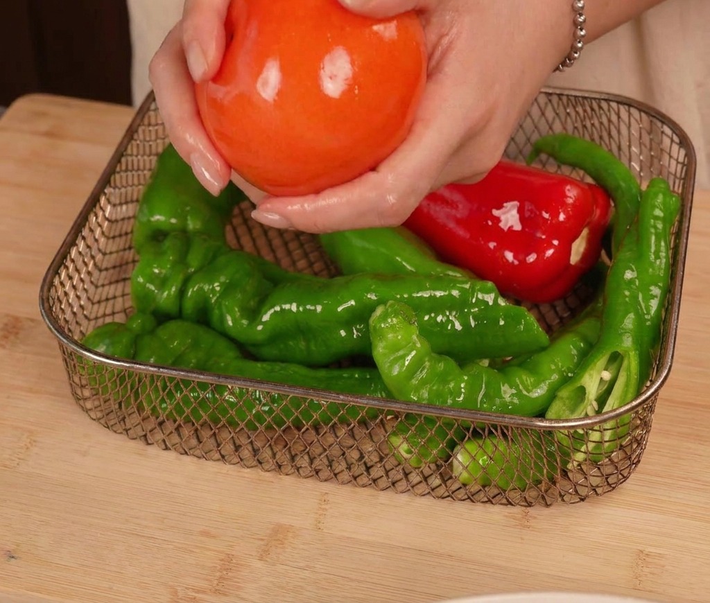 Mani che posizionano un pomodoro lucido e oliato nel cestello della friggitrice ad aria accanto a diversi peperoncini.