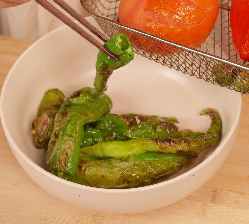 Bacchette che sollevano un peperoncino verde abbrustolito dal cestello della friggitrice ad aria.