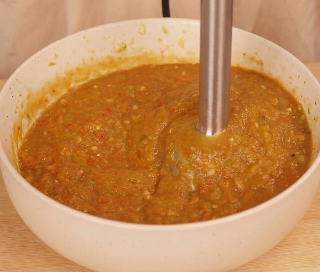 Un frullatore a immersione che trasforma peperoncini e pomodoro in una salsa densa color arancio-marrone.