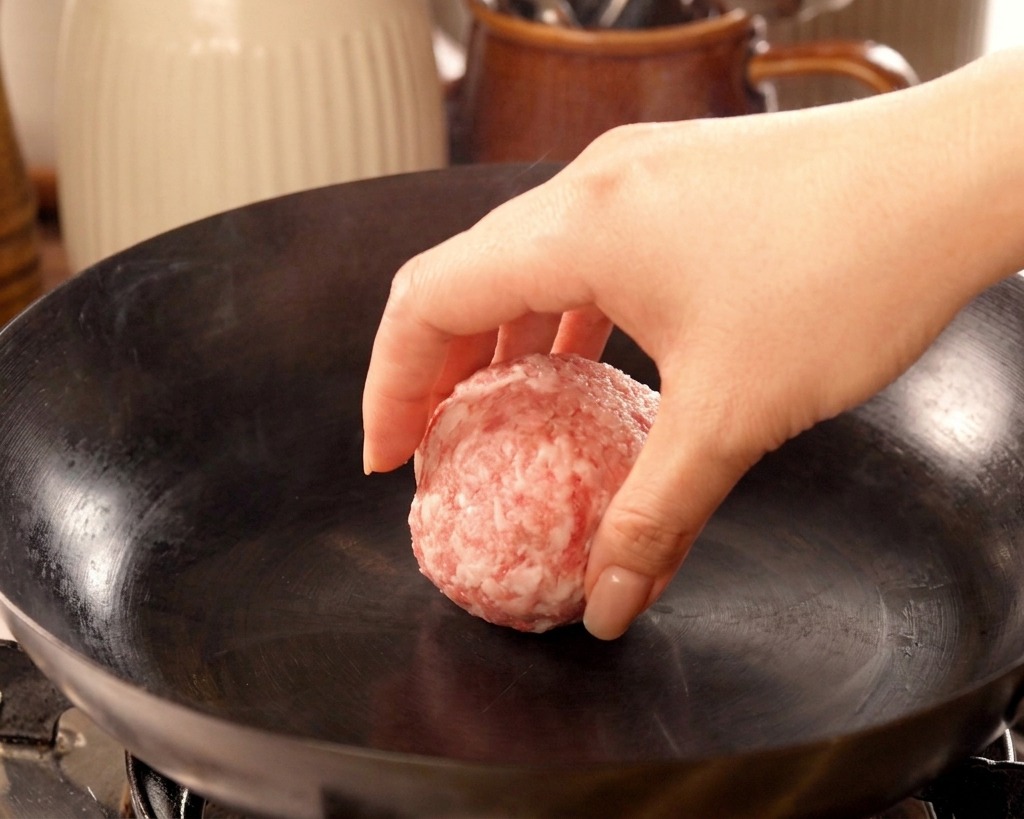 Una mano che adagia una pallina di carne cruda in una padella di ghisa rovente e leggermente fumante.