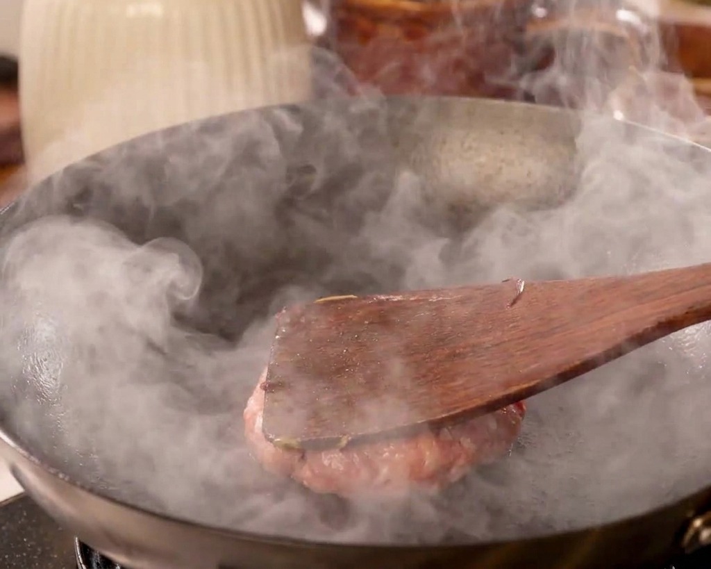 Una spatola di legno piatta che preme con forza su una polpetta di carne appiattendola contro la padella rovente.