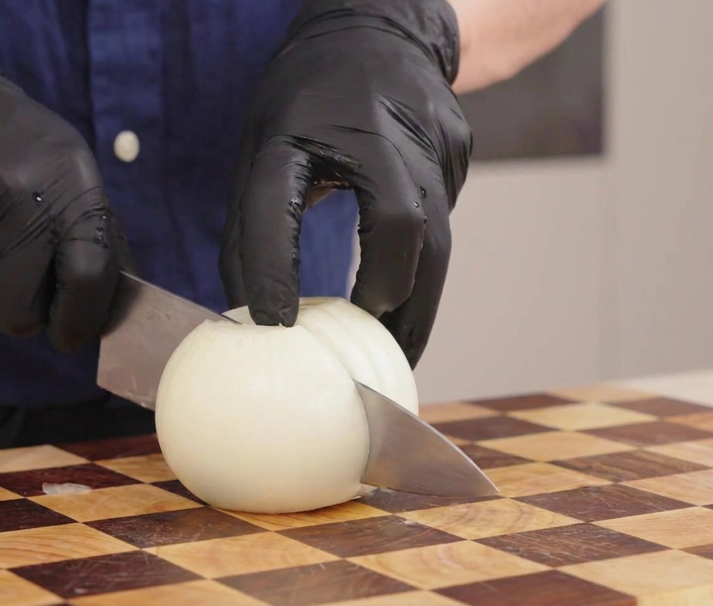 Mani con guanti che usano un coltello per affettare una cipolla bianca a strisce su un tagliere di legno.