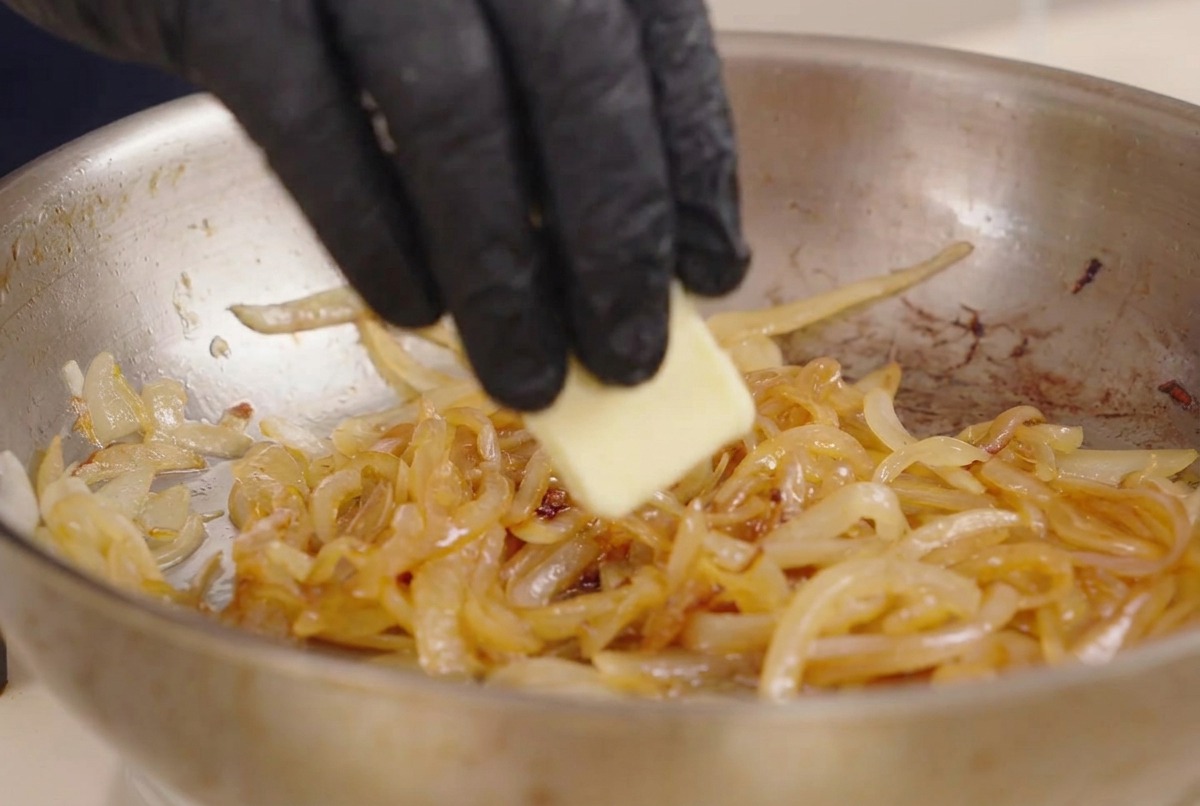 Una mano aggiunge una noce di burro in una padella con cipolle e condimento liquido scuro.