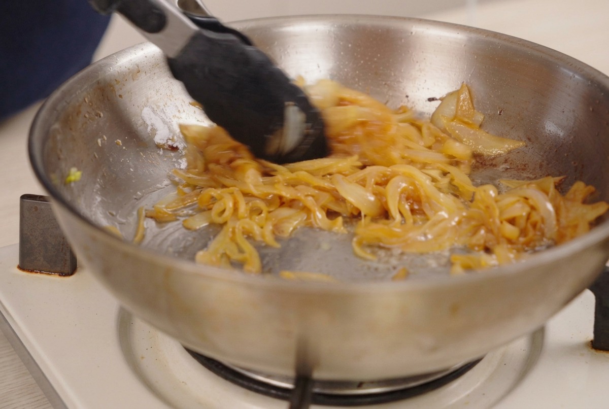 Pinze che mescolano cipolle caramellate marrone scuro in una padella.