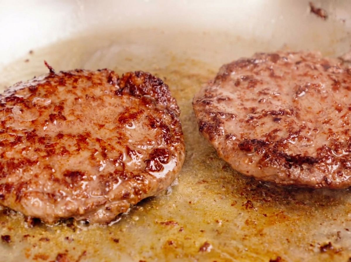 Due polpette di carne spesse che cuociono in padella con una crosticina marrone ben formata.