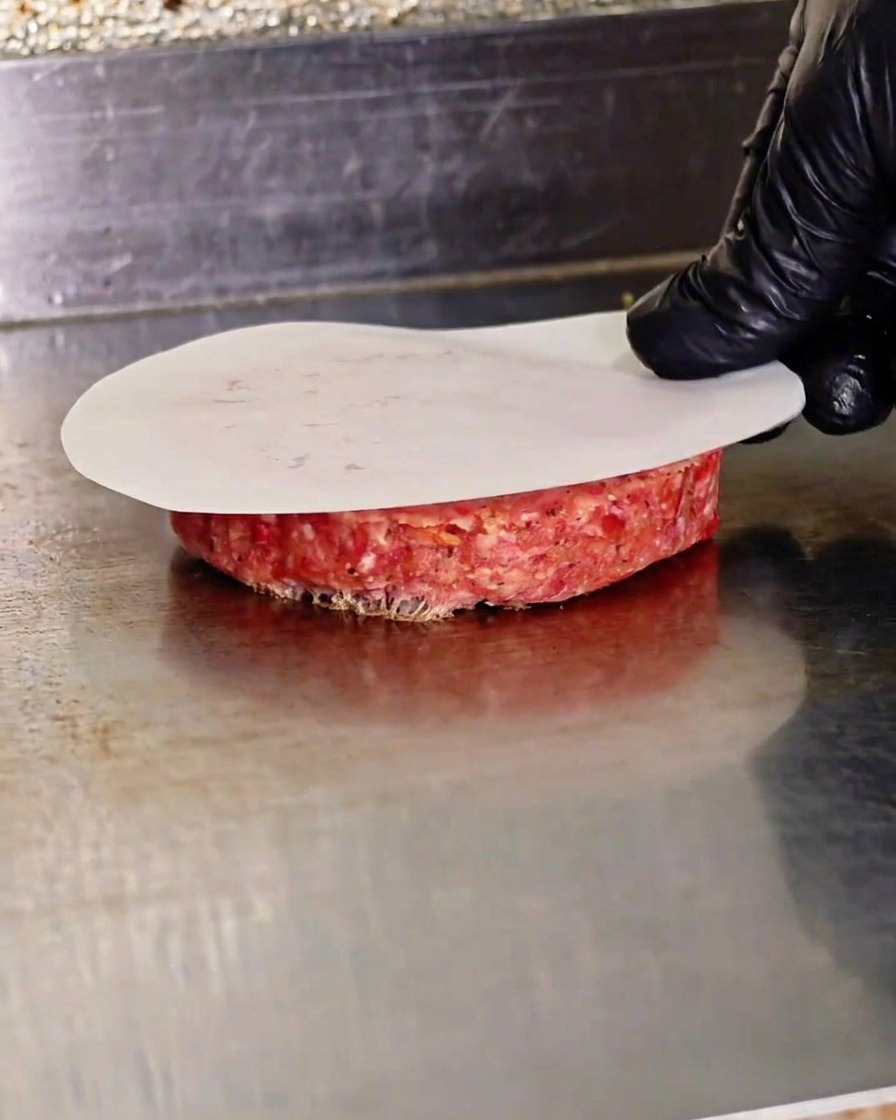 Una mano guantata preme su un patty di carne cruda usando una pressa piatta sopra un foglio di carta forno su una piastra calda.