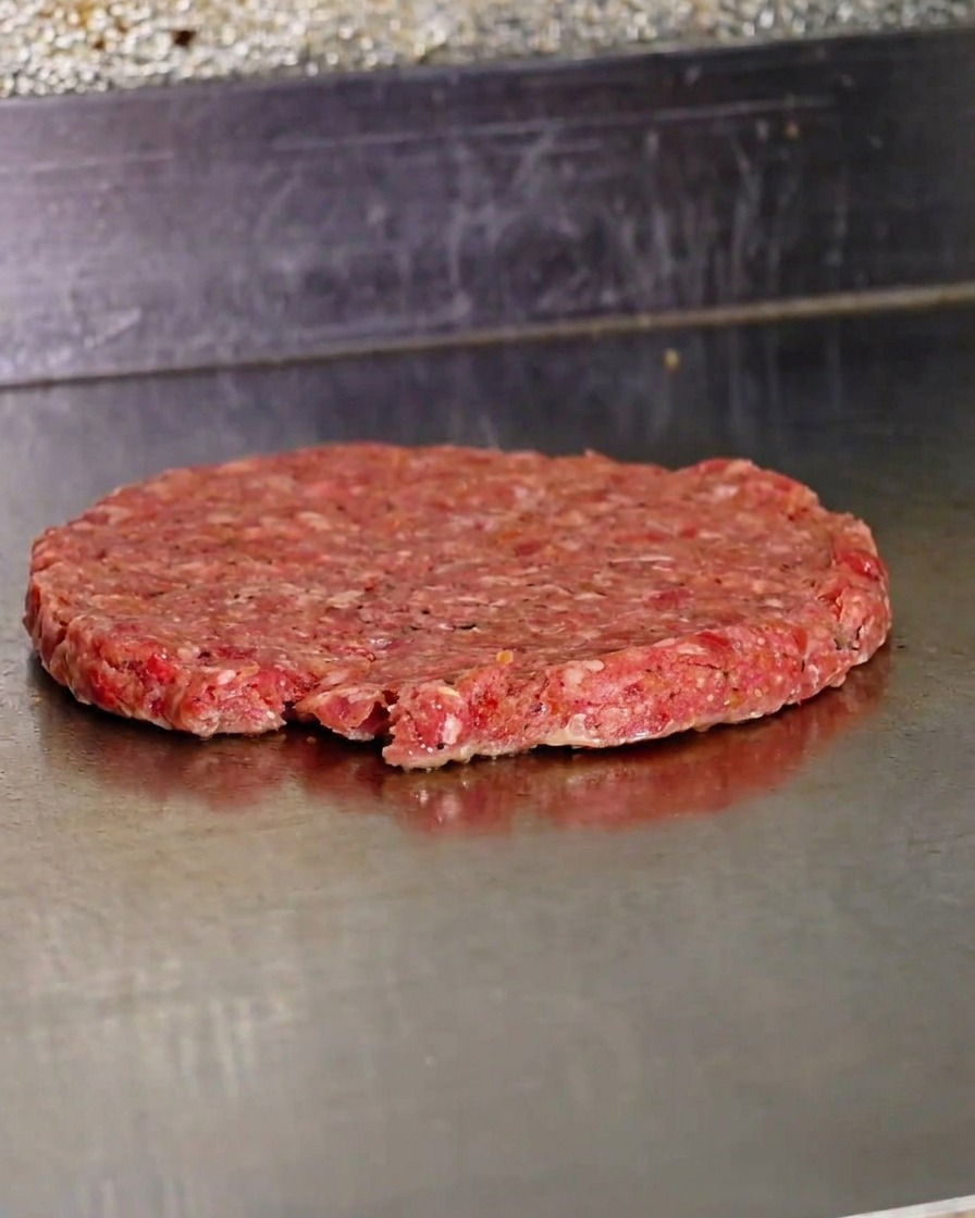 Un patty di manzo appena schiacciato che cuoce su una piastra di metallo rovente.