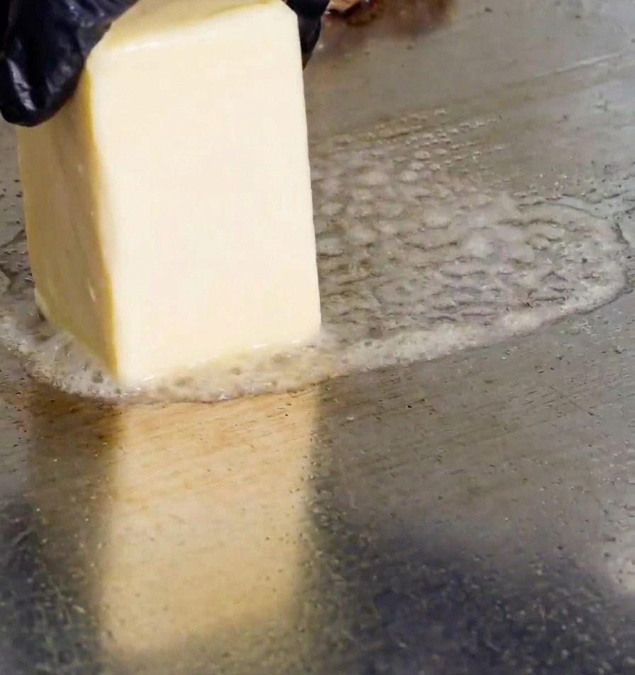 Una mano con guanto nero strofina un panetto di burro su una piastra calda, creando bollicine di grasso fuso.