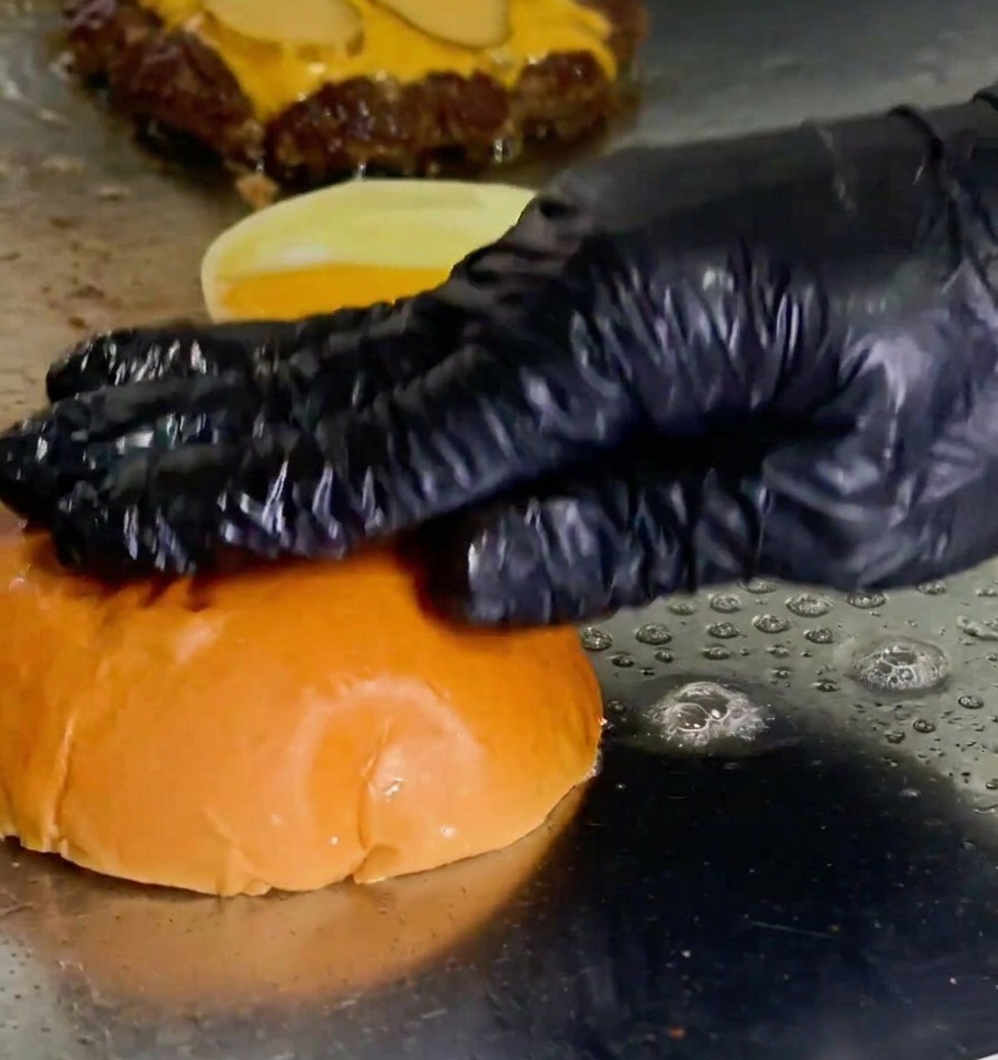 Una mano guantata che preme un bun per burger nel burro fuso sulla piastra accanto a un patty in cottura.