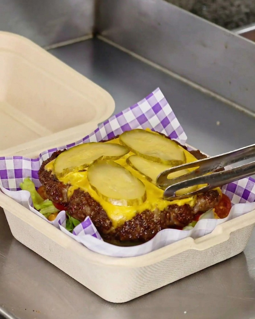 Pinze che trasferiscono un patty smash con formaggio fuso e cetriolini sopra un letto di lattuga e pomodoro.