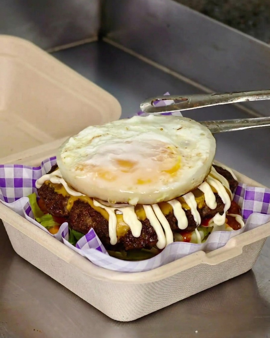 Pinze di metallo che posizionano un uovo fritto sopra un burger ricco di carne, formaggio e maionese.