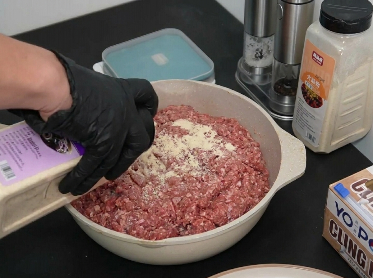 Una mano con guanto spolvera cipolla in polvere sopra una teglia di carne macinata cruda.