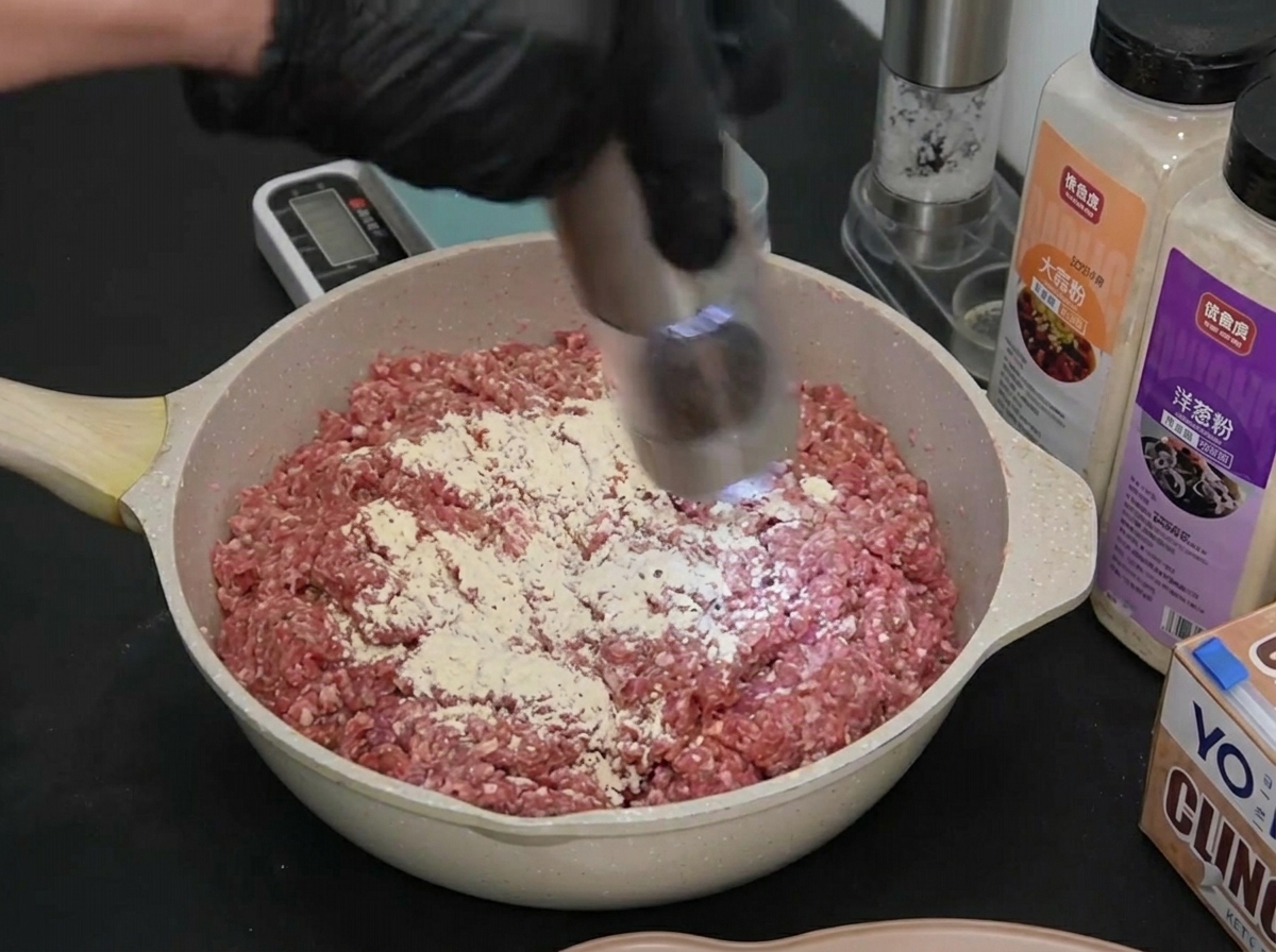 Una mano con guanto usa un macinapepe elettrico sulla carne macinata condita.