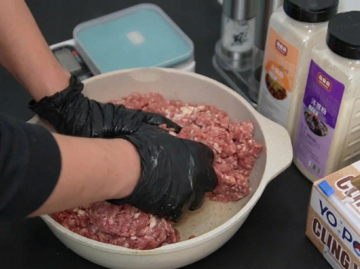 Due mani con guanti premono e mescolano la carne macinata cruda in una teglia.