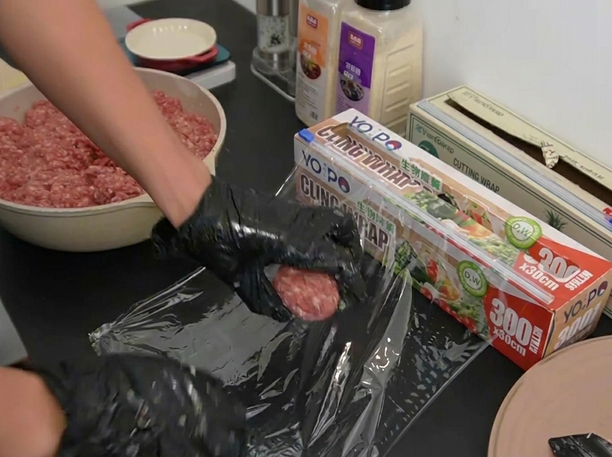 Mani con guanti posizionano una pallina di carne su un foglio di pellicola trasparente.