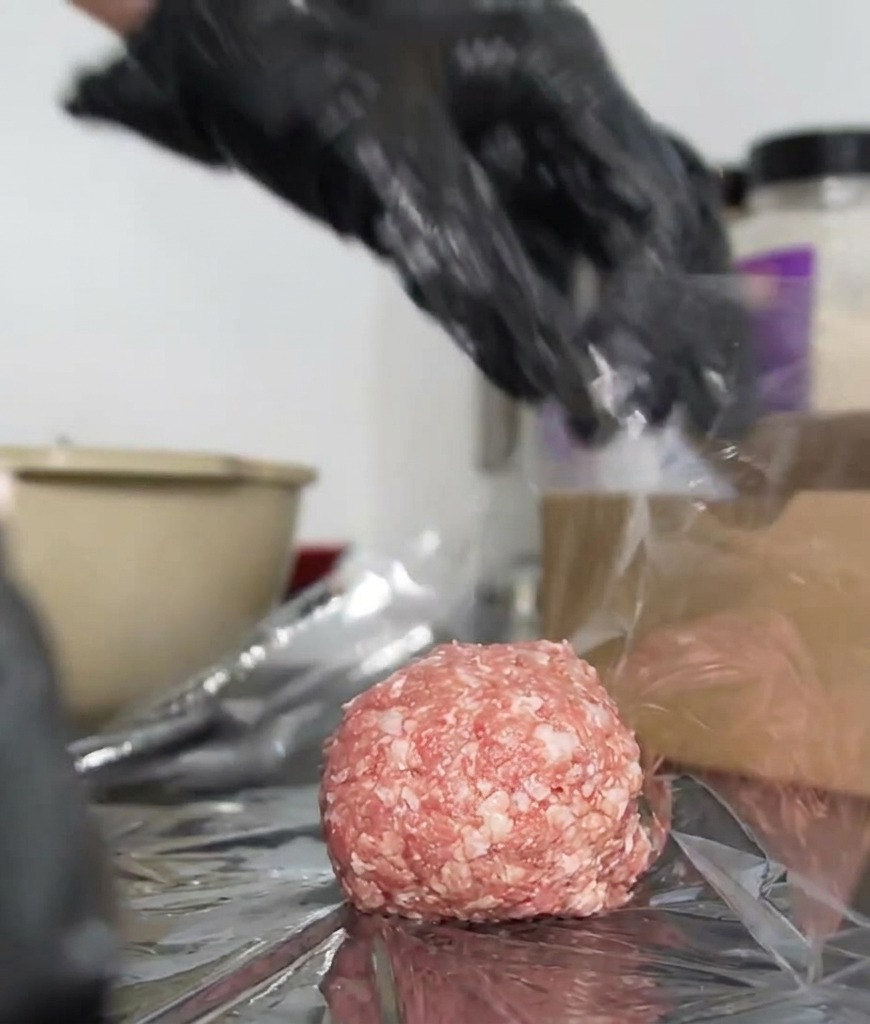 Mani con guanti che ruotano la pellicola trasparente attorno a un hamburger per sigillarlo.