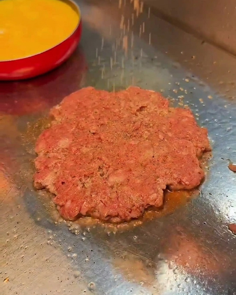 Sale e pepe nero sparsi su un patty di manzo che cuoce sulla piastra.