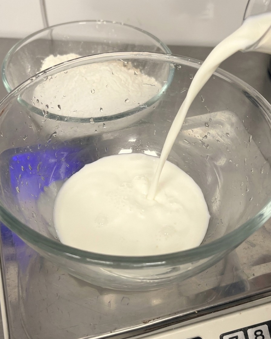 Latte fresco versato in una ciotola di vetro trasparente con farina sullo sfondo.