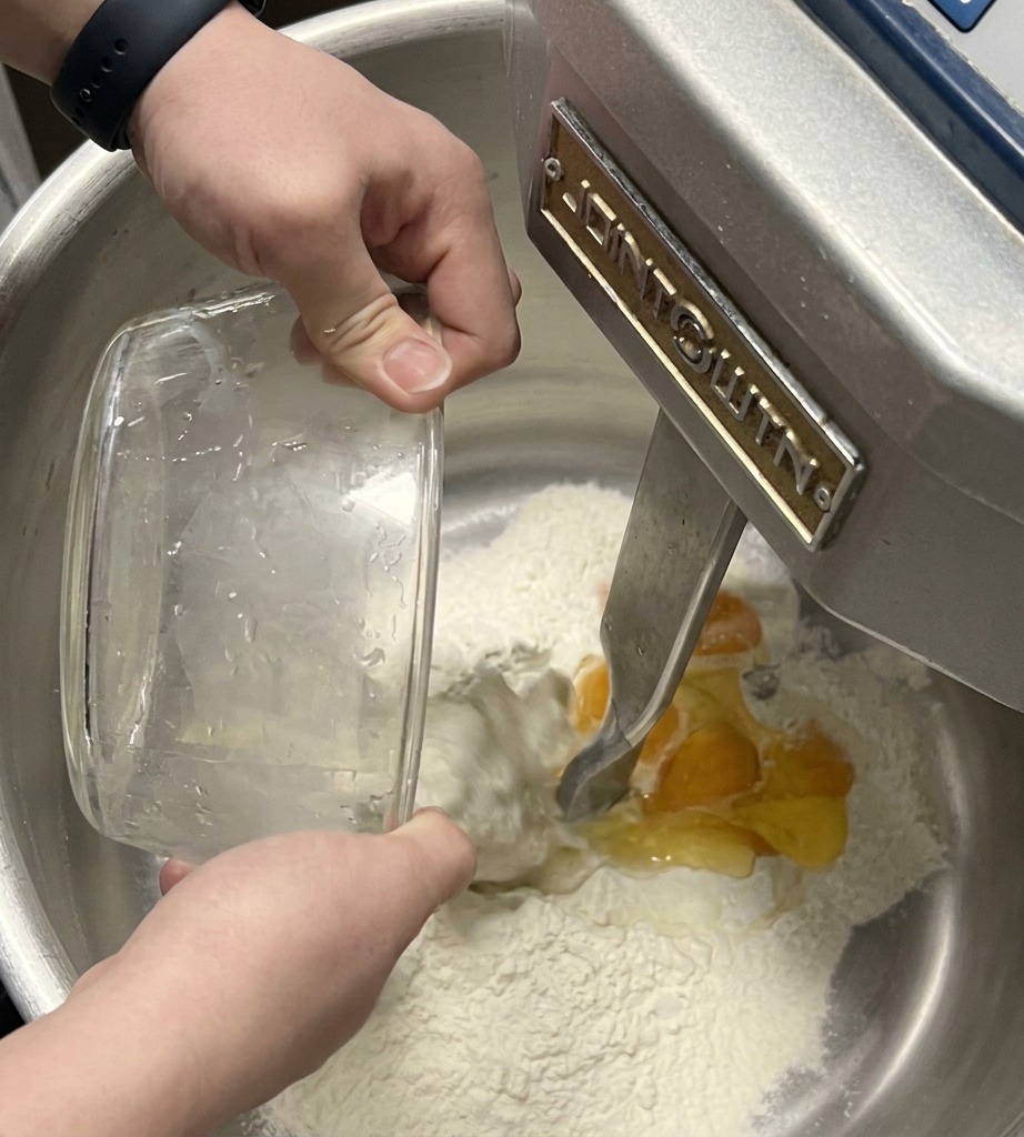 Gancio di unimpastatrice che incorpora uova e liquidi nella farina in una ciotola dacciaio.
