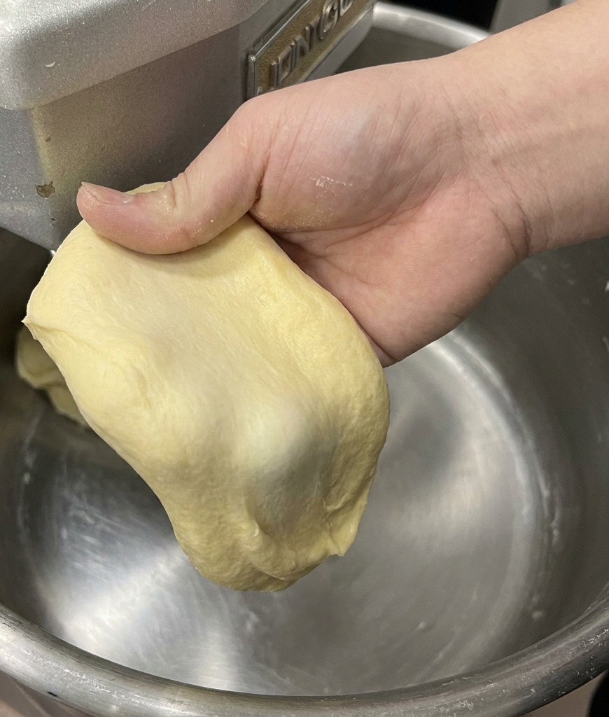 Una mano che tende un pezzetto di impasto brioche giallo chiaro per mostrare la membrana sottile e resistente.