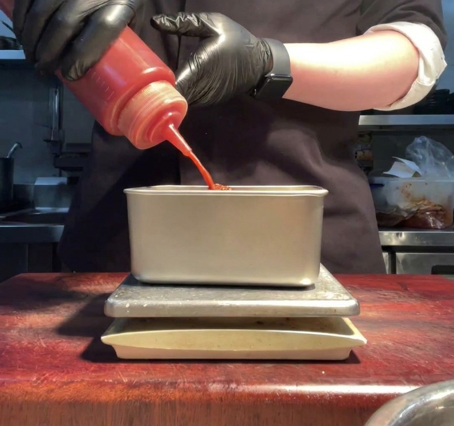 Mani guantate che spremono salsa sriracha rossa in un contenitore di metallo con maionese.