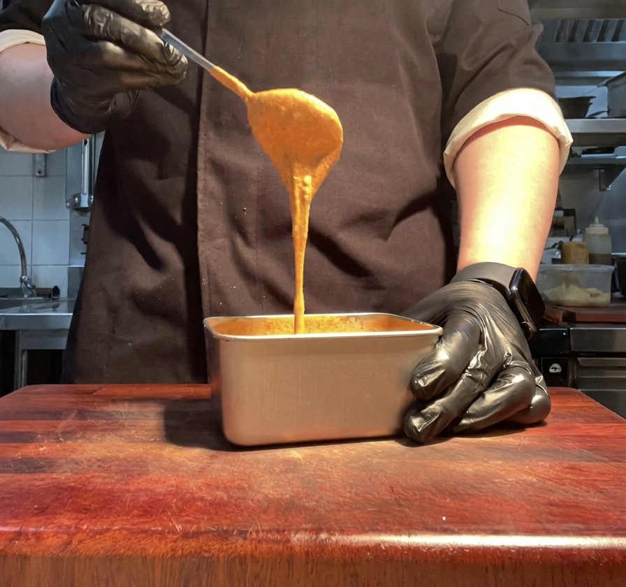 Una mano guantata che solleva un cucchiaio di salsa arancione vibrante da un contenitore metallico.