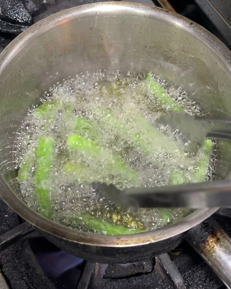 Pinze metalliche che muovono peperoncini verdi interi mentre friggono in olio bollente.