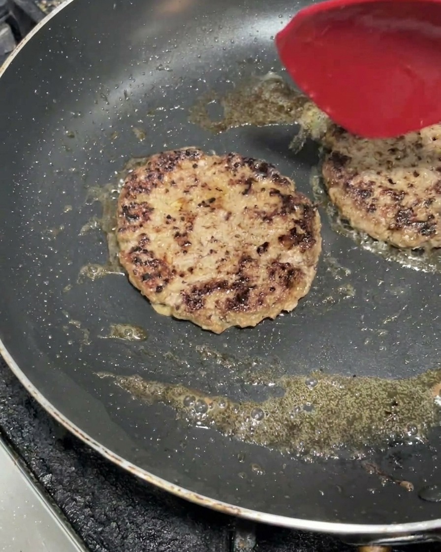Due polpette di manzo spesse che sfrigolano in padella con una crosta ben dorata e una spatola rossa.
