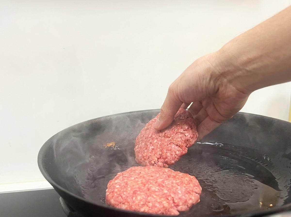 Una mano che adagia due polpette di manzo in una padella nera rovente e leggermente fumante.