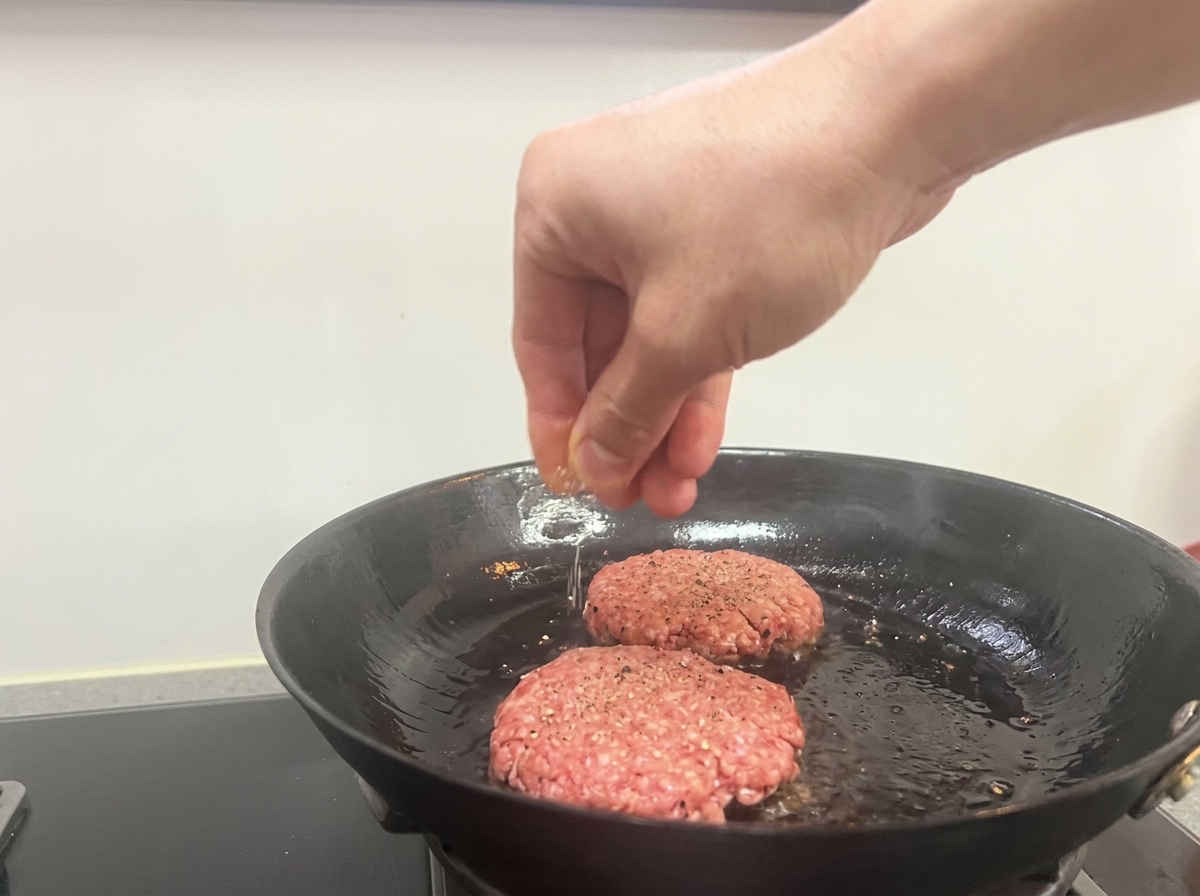 Una mano che cosparge sale e pepe nero su due patty di manzo che sfrigolano in padella.