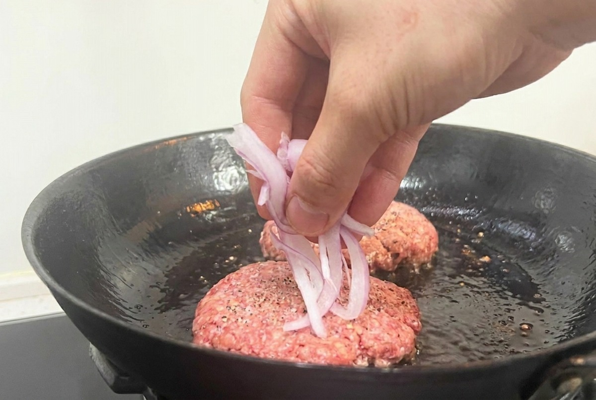 Una mano che sparge fettine di cipolla rossa sui patty di carne cruda in una padella nera.