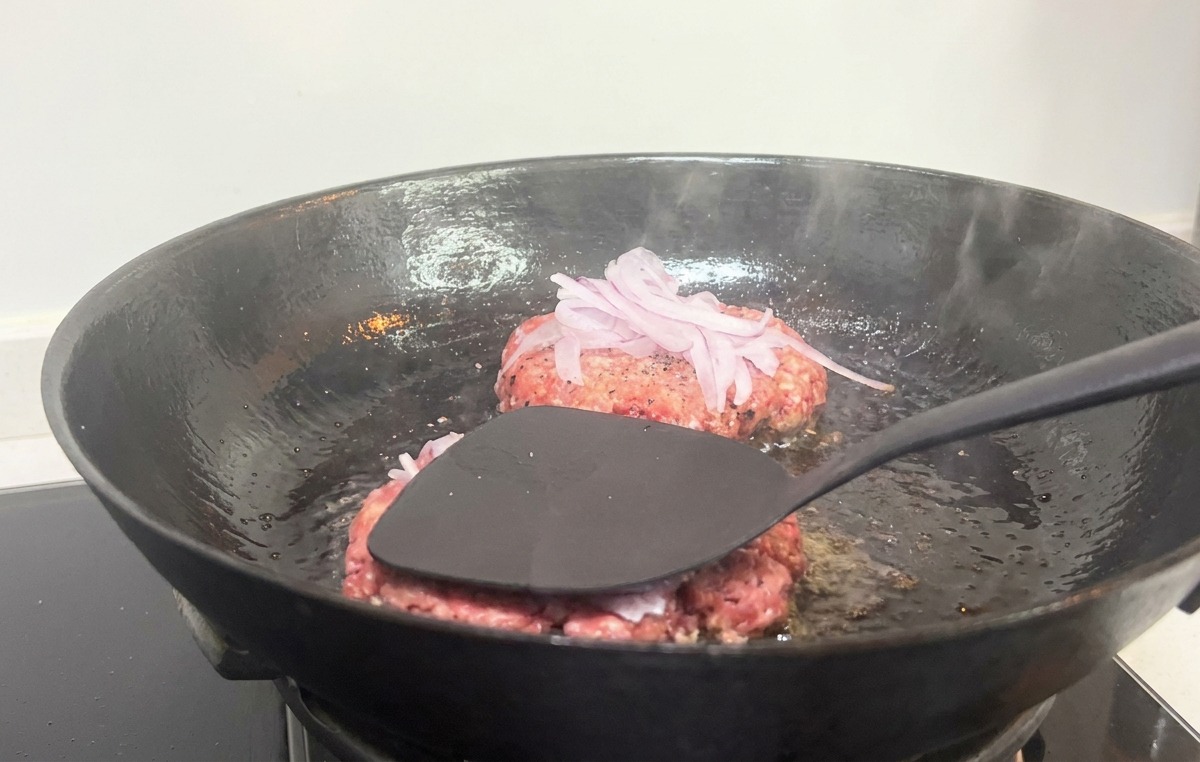 Una spatola nera che preme con forza su un burger con cipolle rosse in una padella di ghisa calda.