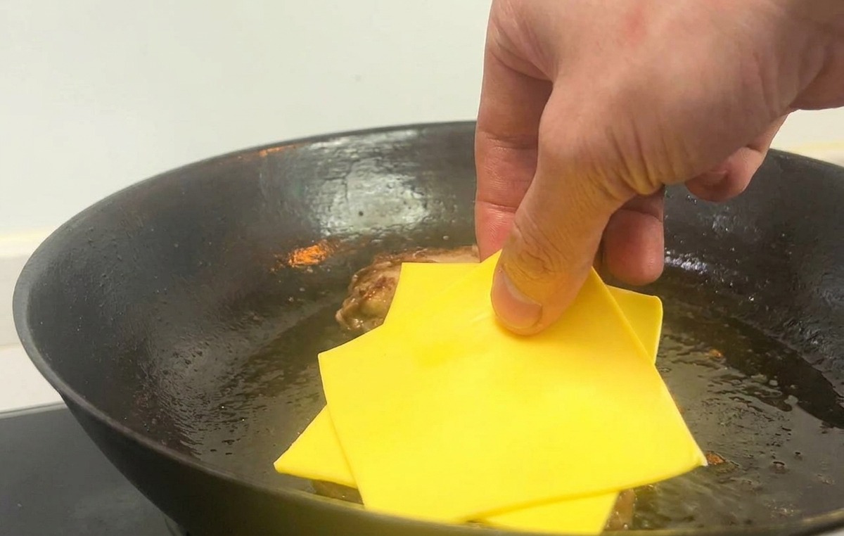 Una mano che mette due fette quadrate di formaggio giallo sui patty cotti in padella.